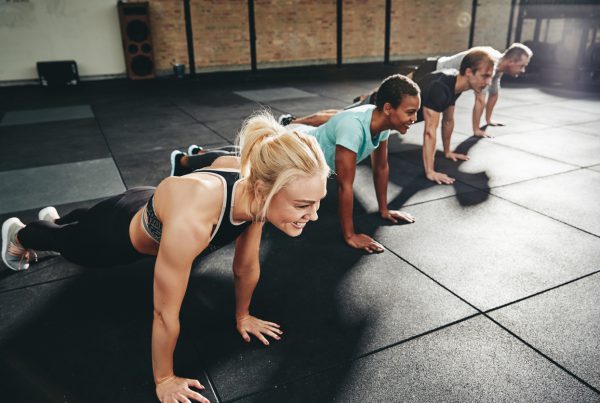 Diverse group of fit people in sportswear smiling while doing pushups together on a gym floor during an exercise session