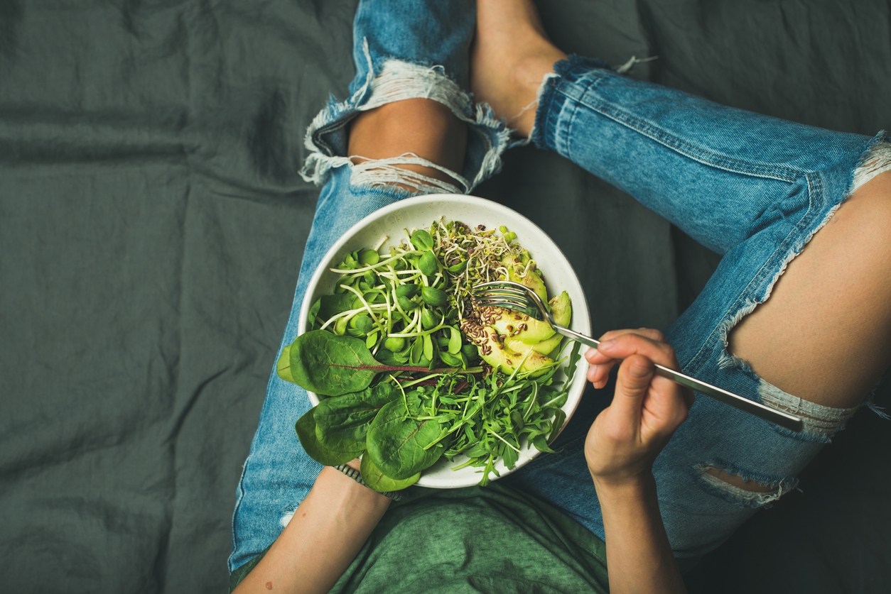 Plant-based eating - Image of green vegan breakfast meal in bowl with spinach, arugula, avocado, seeds and sprouts. Girl in jeans holding fork with knees and hands visible