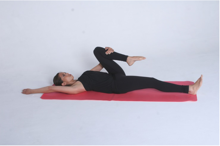 Image of Hip Extension Assessment - this shows a woman laying on her back on a at with one leg straight out on the floor whilst the other leg is bent with her hand holding her knee in towards her body