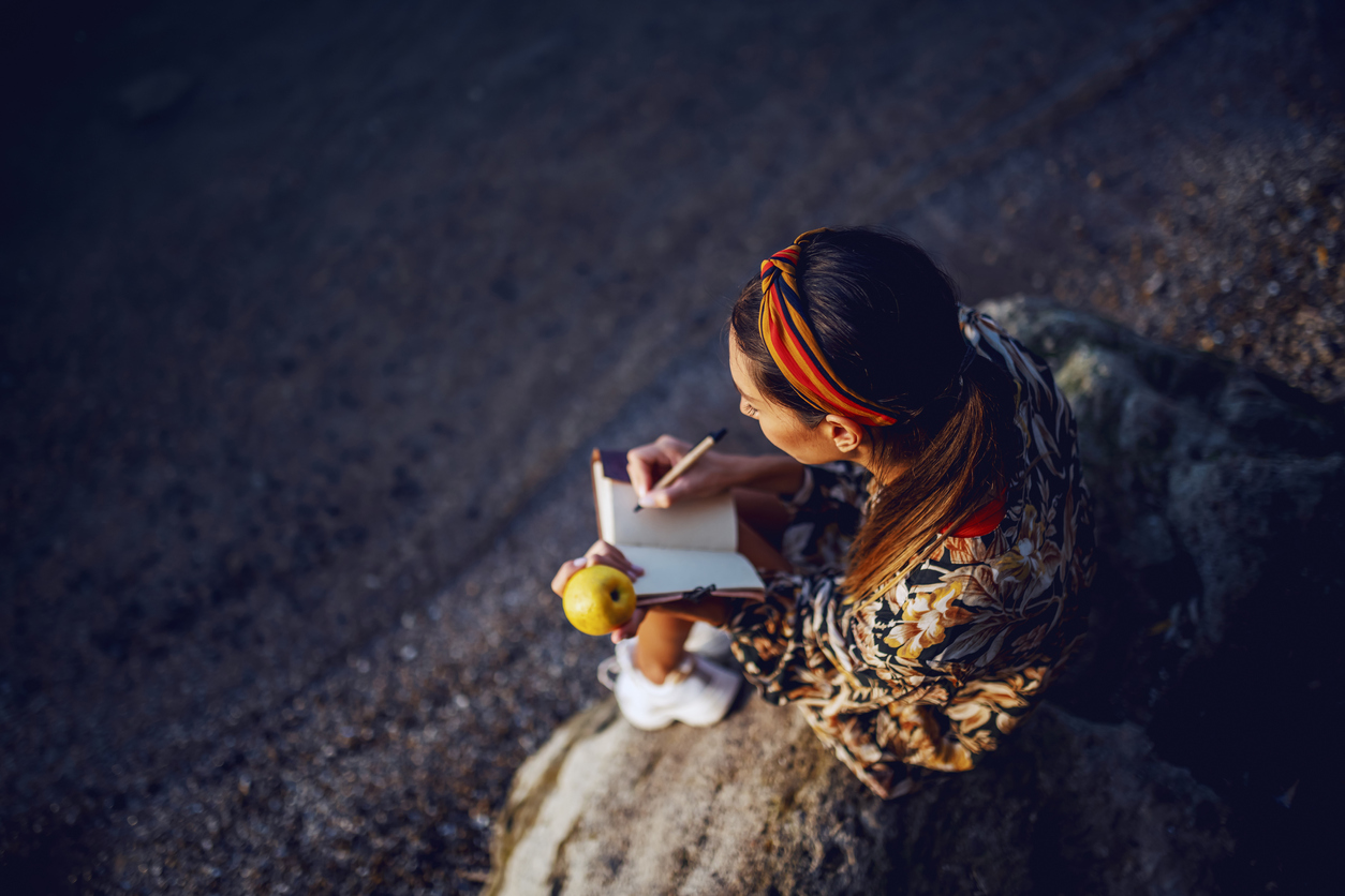 Mindfulness - Image of top view of woman in floral dress and with headband sitting on the rock on shore, eating apple and writing diary.