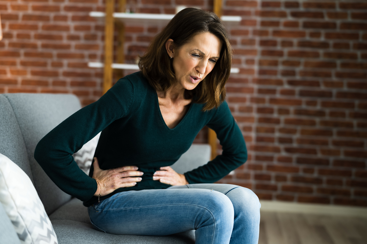 Endometriosis - Image of woman clutching her lower abdomen looking in pain. She is sat on a sofa with brick wall in the background.