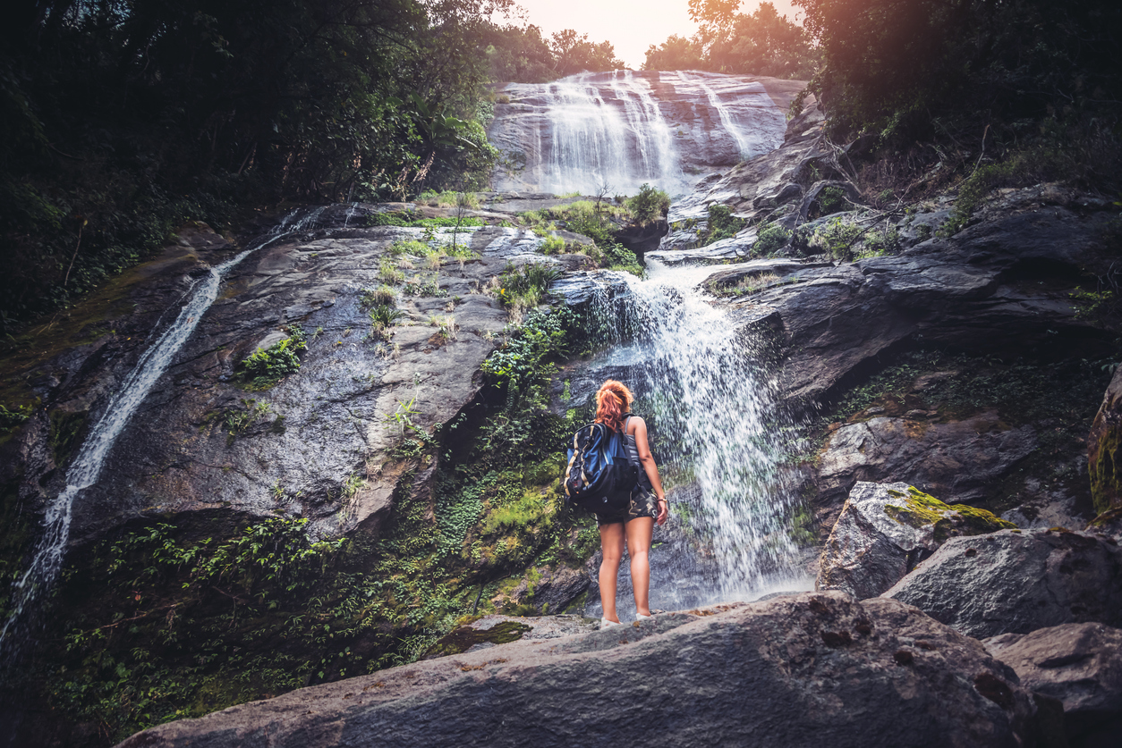 Blue Space. Image of woman looking up at a waterfall with a ruck sack on her back. It appears she is on a walking trail taking a moment to take in the splendour of the waterfall.