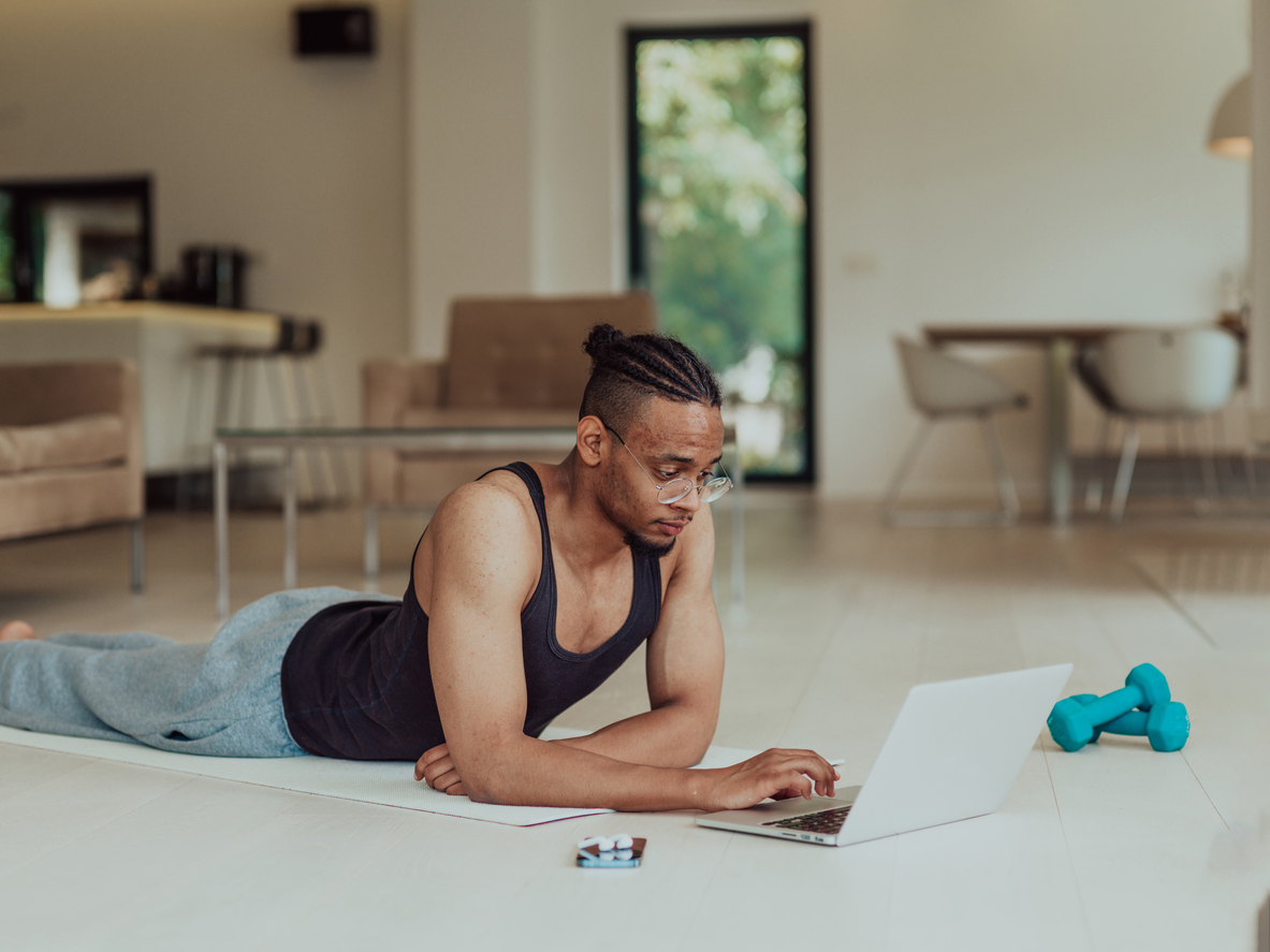 Tips for Instructors and PTs. Image of man who wears glasses is at home looking at his laptop whilst lying on a yoga mat. It looks like he is studying. He has got a set of dumbbells near by and a phone and headphones.