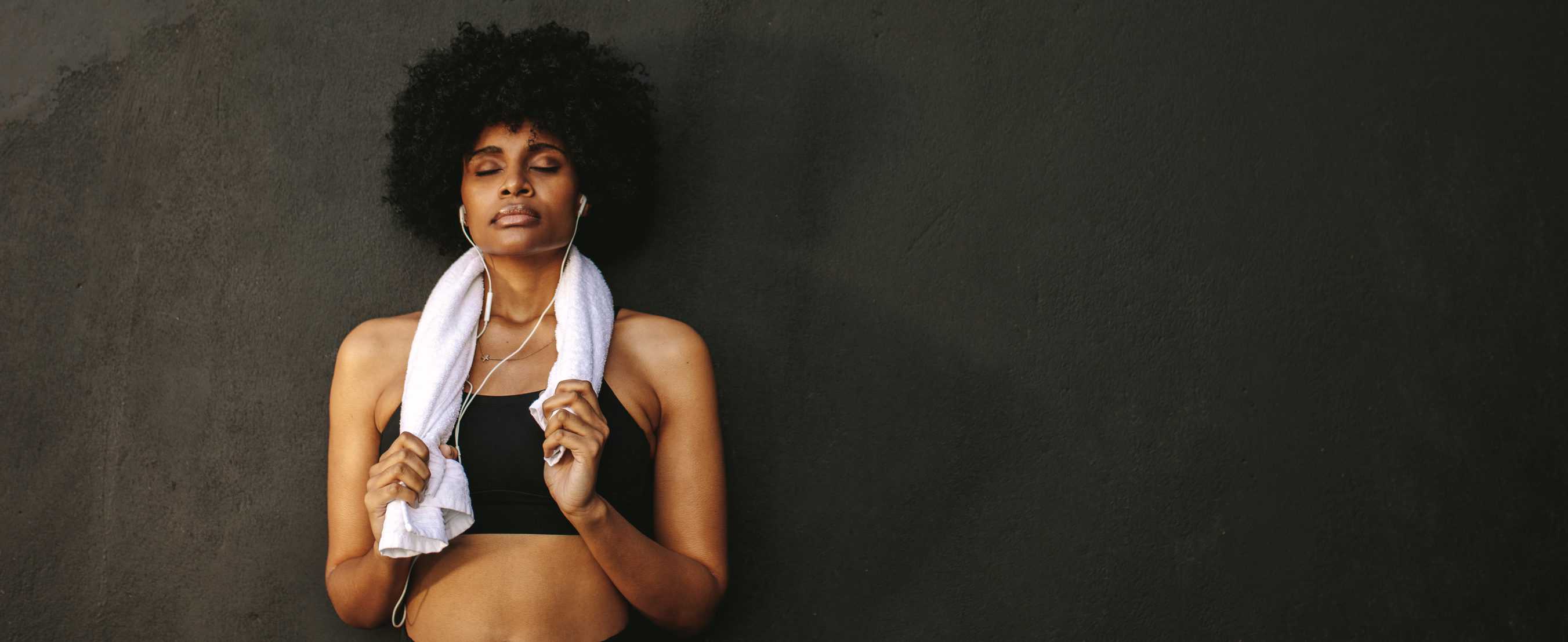Struggle of group exercise - Image of female athlete resting after workout with eyes closed leaning against a wall. She looks tired with a towel hanging around her neck.