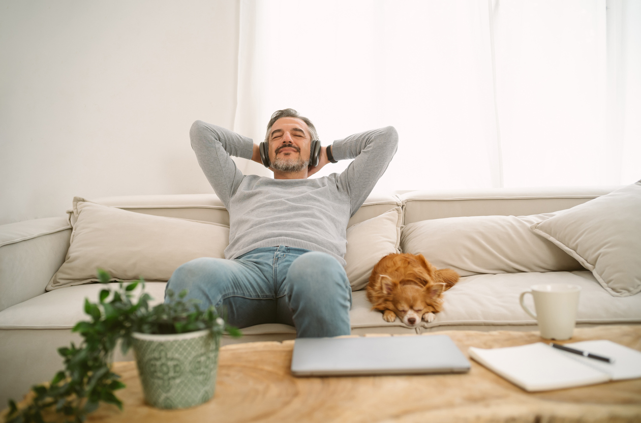 Recover - Image of a relaxed man wearing wireless headphones, leaning back on a sofa with his content Chihuahua lying next to him asleep